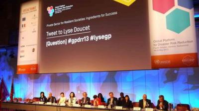 A panel of speakers sits at a long table on stage at the Global Platform for Disaster Risk Reduction, with a large screen displaying event information behind them.