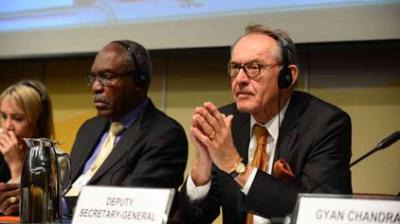 Three people sit at a conference table wearing headsets; the man in front is labeled as the Deputy Secretary-General.