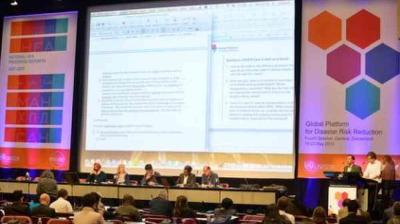 A conference panel sits at a long table facing an audience, with large presentation slides projected on screens behind them.