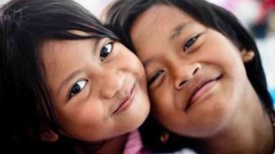 Two children smiling closely together, with their heads touching and looking directly at the camera.