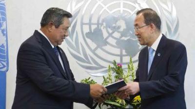 Two men in suits exchange a document in front of a United Nations emblem, with flowers and a flag in the background.