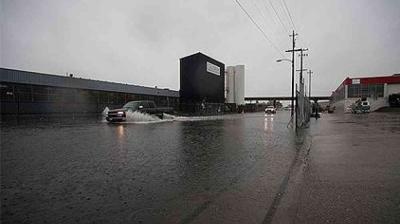 Vehicles drive through a flooded industrial street on a rainy day, with water covering most of the road and overcast skies overhead.