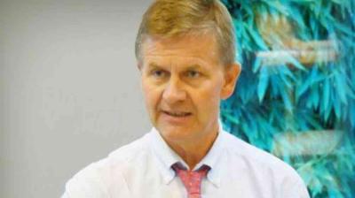 A man in a white shirt and red tie is speaking indoors, with a blurred, leafy background behind him.
