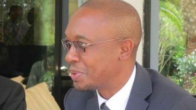 A man in a suit and glasses sits indoors, facing left, with greenery visible outside through a window behind him.