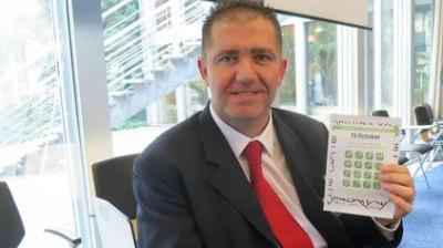 A man in a suit and red tie sits indoors, holding up a calendar or planner open to the month of October.