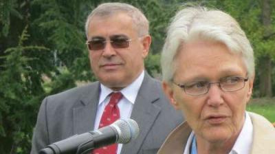 Two older adults, a man in a suit and a woman with glasses, stand outdoors near a microphone with trees in the background.