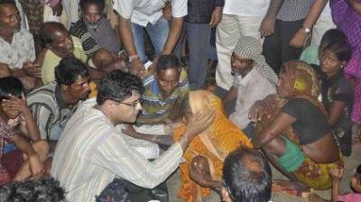 A man in glasses talks to and comforts a woman in a yellow sari, surrounded by a group of seated and standing people in an indoor setting.