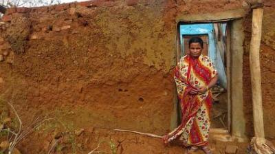 A woman in a patterned sari stands in the doorway of a partially collapsed mud brick house.