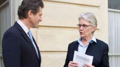 Two people in business attire stand outside a building, having a conversation; one holds printed documents.