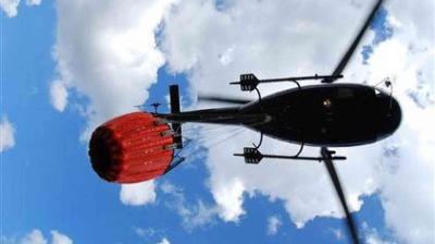 A helicopter carrying a large red water bucket flies overhead against a blue sky with scattered clouds.