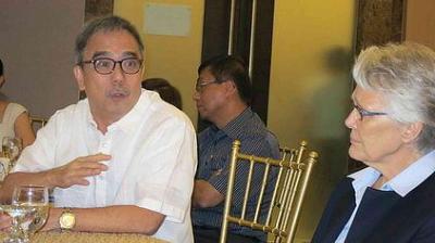 Two people are seated at a table, engaged in conversation during an indoor event. Other attendees are present in the background.