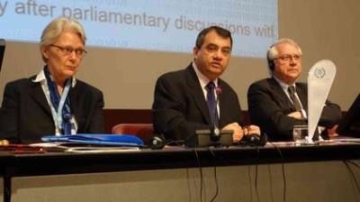 Three people in formal attire sit at a conference table with microphones, laptops, and documents, speaking at a meeting or panel discussion.