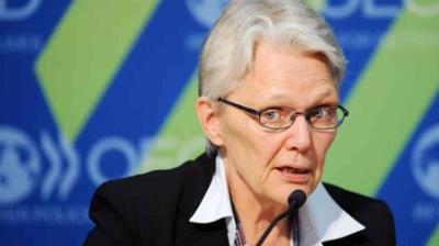 An older person with short gray hair and glasses speaks into a microphone, wearing a suit jacket and white collared shirt, with a blue and green background behind them.