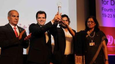 Four people in formal attire stand on stage; one man in the center holds up a trophy while others applaud during an award ceremony.
