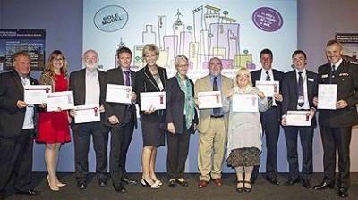 A group of eleven people stand in a row holding certificates, posing for a photo in front of a colorful illustrated backdrop at an event.