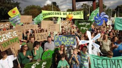 A large crowd holds signs demanding climate action and clean energy at an outdoor protest; banners include “FUTURE IS NOW” and “CHANGEONS DE RÉGIME!”.