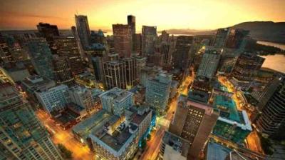 Aerial view of a city skyline at sunset, with tall buildings, illuminated streets, and a body of water in the background.
