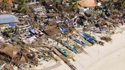 Aerial view of a coastal area with damaged houses, scattered debris, and boats on a sandy beach, likely after a natural disaster.