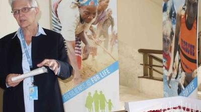 A person stands indoors holding a folder, with banners featuring people and the text "RESILIENCY IS FOR LIFE" displayed in the background.