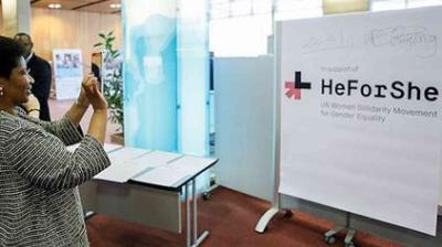 A person takes a photo of a large "HeForShe" sign at an indoor event promoting UN Women's solidarity movement for gender equality.