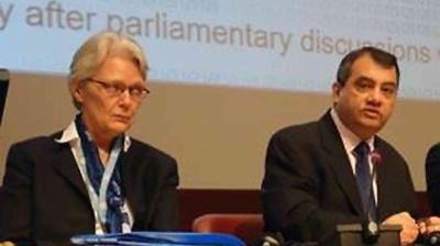 Two people in business attire are seated at a conference table, speaking in front of a projection screen with partial text visible in the background.