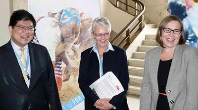 Three adults in business attire stand indoors, smiling and conversing, with a staircase and a large poster featuring children in the background.
