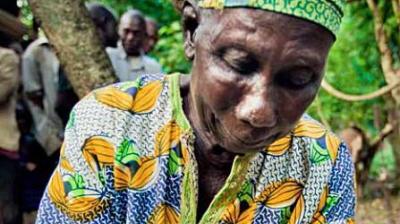 An older person wearing a patterned shirt and headscarf looks down while outdoors, with several people and trees visible in the background.