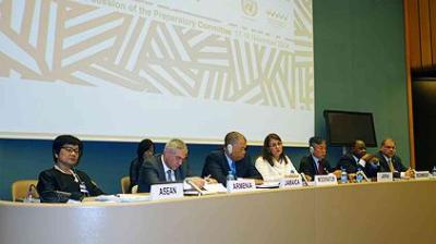 A group of people are seated at a conference table with nameplates for ASEAN, Armenia, Jamaica, Morocco, Japan, and others. A large screen displays text related to a UN conference.