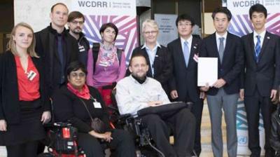 Group of individuals, including a person in a wheelchair, standing together indoors at the WCDRR event. Various banners are visible in the background.