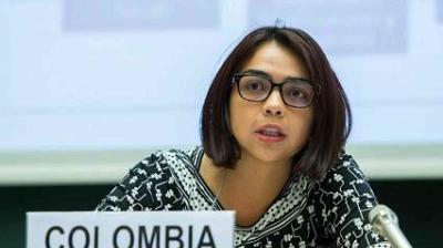 A woman wearing glasses speaks into a microphone at a conference, seated behind a sign that reads "COLOMBIA.