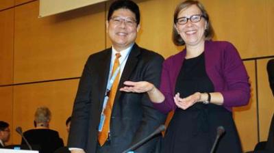 Two people, one man in a suit and tie and one woman in a dress and purple cardigan, stand smiling at a conference table with microphones in front of them.