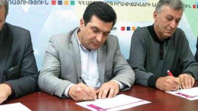 Three men sit at a table, signing documents with pens. A colorful banner with Armenian text is visible in the background.