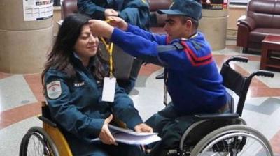 A man in a wheelchair places a lanyard around a woman's neck; she is also in a wheelchair. They appear to be in a public indoor space.