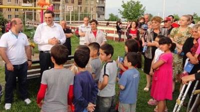 A group of adults and children gather outdoors, some standing in a semi-circle, while one man in a white shirt appears to address the group.