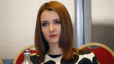 A woman with straight, shoulder-length brown hair sits in front of red chairs, wearing a black and white patterned top. A bottle is partially visible in the foreground.