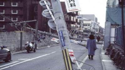 A street with fallen utility poles, damaged buildings, and debris; a person walks alongside.