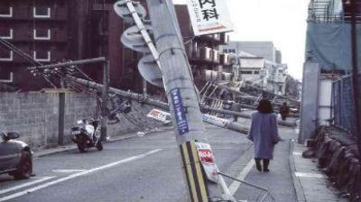 A street with multiple fallen utility poles leaning across the road; a person walks along the sidewalk beside the damage.