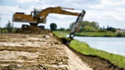 An excavator works on leveling or moving soil near a body of water, with a dirt embankment in the foreground and greenery in the background.