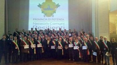 A group of people in formal attire stands on a stage holding certificates, in front of a screen that reads "Provincia di Potenza, Network Comuni Resilienti.