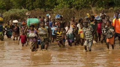 A large group of people wade through muddy floodwaters, carrying belongings on their heads and in their hands, surrounded by dense vegetation.