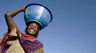A smiling person wearing a colorful garment balances a large blue basin on their head against a clear blue sky.