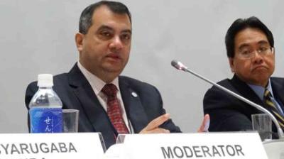 Two men in formal attire seated at a conference table with microphones. The man on the left speaks while the man on the right listens. A water bottle and name cards are visible.