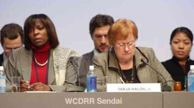 A group of people seated at a conference table with nameplates and water bottles; the nameplate in focus reads "TARJA HALONEN" and the backdrop says "WCDRR Sendai.