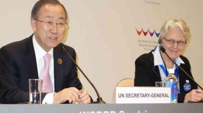 Two individuals seated at a conference table, one speaking at a microphone labeled "UN Secretary-General" and the other listening attentively.