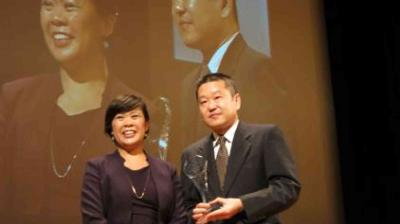 Two people in formal attire stand on stage, one holding a trophy. A projected image of them is visible in the background.