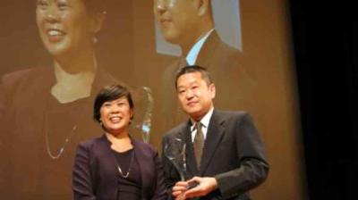 Two people in formal attire stand together on stage holding an award, with their image projected on a large screen behind them.