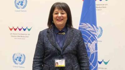 A woman in a dark suit stands in front of United Nations banners, smiling at the camera with an ID badge visible on her chest.