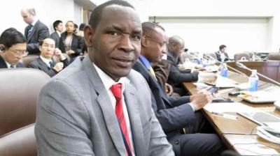 A man in a gray suit and red tie sits at a conference table with other attendees, some using laptops and tablets, in a formal meeting room.
