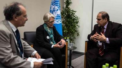 Three people in formal attire have a discussion in an office setting with a UN flag and water bottles visible in the background.