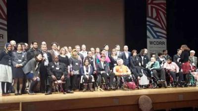A large group of people, including several in wheelchairs, pose together on stage at an event with banners labeled "WCDRR" in the background.